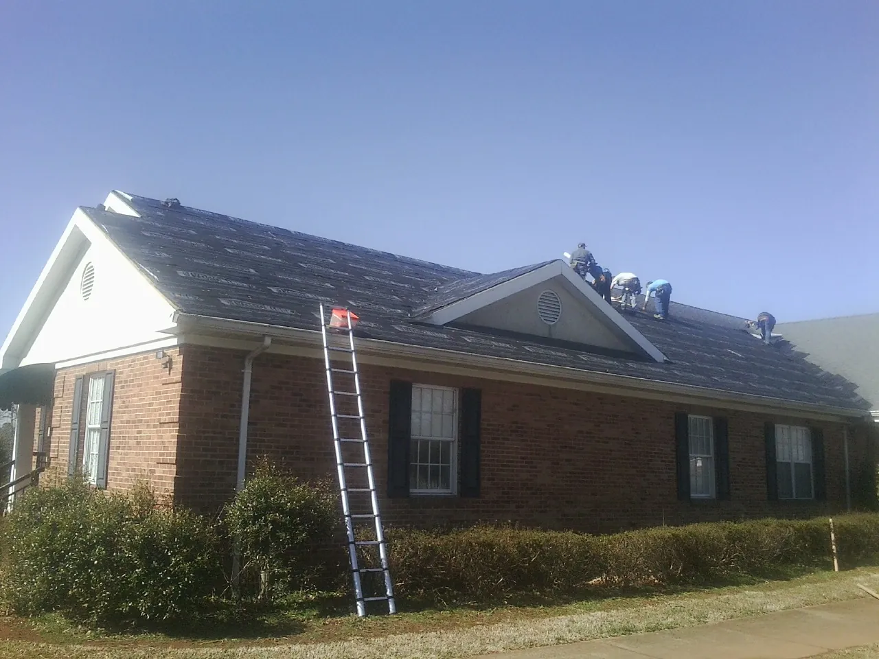 Crew tearing shingles off a brick ranch roof with a ladder set — Emergency Roof Repair work by our crew in Copiague