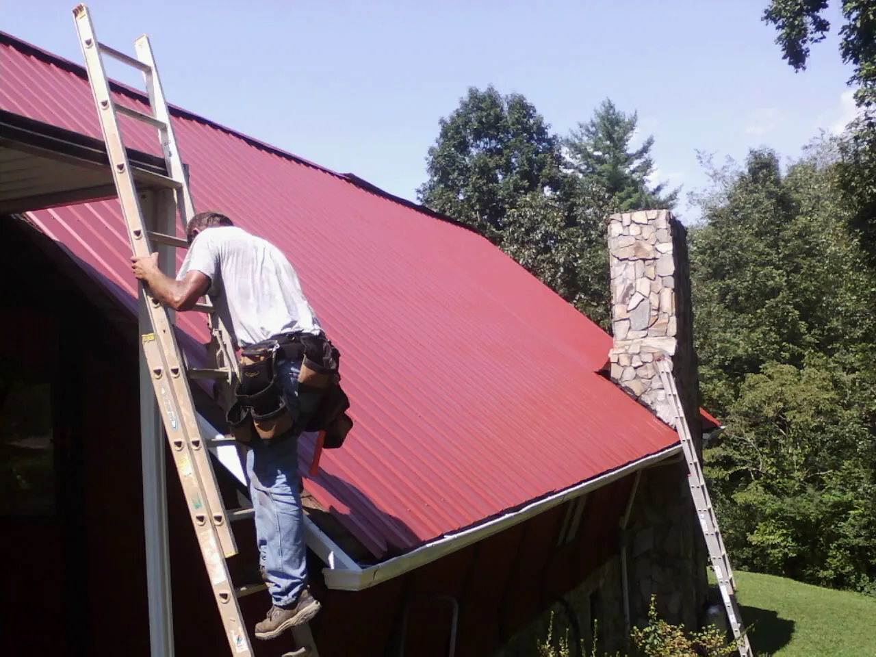 Roofer on a ladder beside a red metal roof and stone chimney — Soffit & Fascia Repair by Roofing Brothers in Copiague